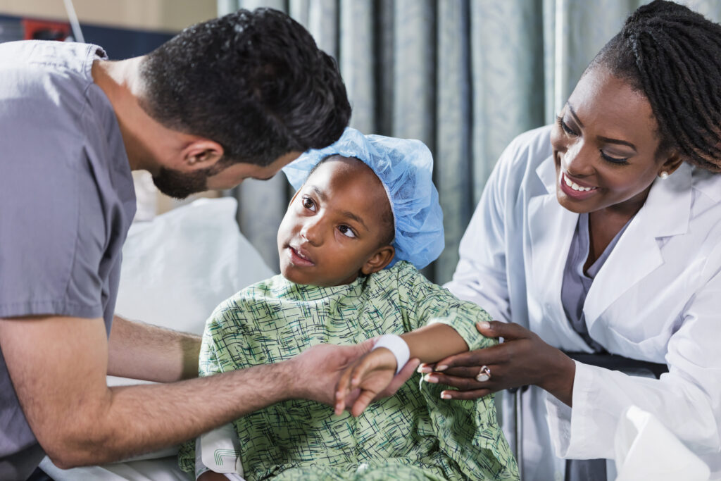 Une fille de 6 ans d’origine afro-américaine est assise sur un lit d’hôpital. Une médecin et un infirmier s’occupent d’elle.  Elle porte une blouse d’hôpital et une charlotte chirurgicale. Un infirmier en tenue médicale, un jeune homme dans la vingtaine originaire du Moyen-Orient, se tient d’un côté et une médecin, une femme dans la quarantaine d’origine afro-américaine, se tient de l’autre côté.