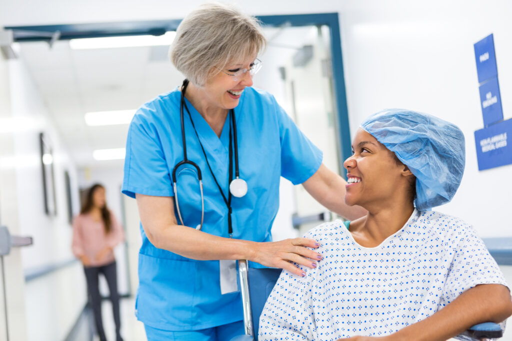 Infirmière caucasienne adulte âgée aux cheveux gris pousse un patient en fauteuil roulant. Elle emmène le patient en salle de réveil après une intervention chirurgicale ambulatoire. Le patient est une jeune femme afro-américaine portant une blouse d'hôpital et une charlotte chirurgicale. Les femmes sourient en se parlant.